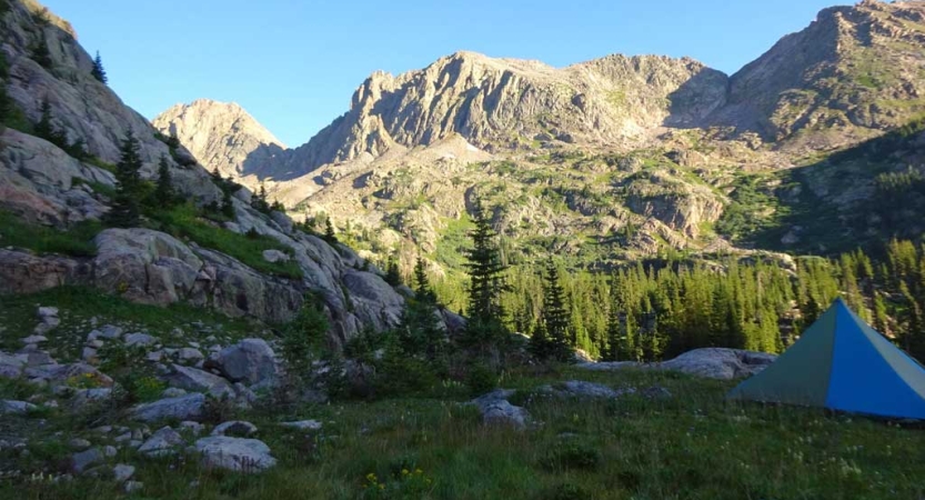 A tent rests in a green meadow amid tall rocky mountains